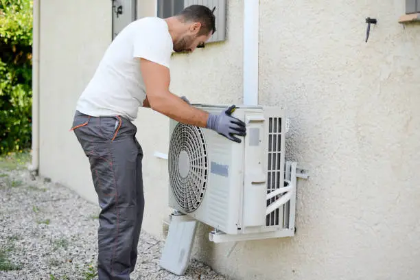 Technician replacing outdoor AC condenser beside occupied Hempstead home in summer